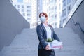 Unhappy fired masked woman is standing in the street with a box of personal items from the desktop. An unemployed