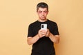 Unhappy injured sufferer man in black T-shirt with bruises and abrasions on his face isolated over beige background using mobile