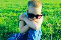 Unhappy little boy wearing sunglasses sitting on green grass. Cute little boy outdoors. Royalty Free Stock Photo