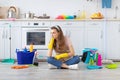 Unhappy millennial housemaid sitting on kitchen floor with heaps of supplies, sick and tired of cleaning