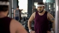 Unhappy overweight man looking at his mirror reflection in gym, diet and sport