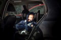 Unhappy, sad child sitting in a child car seat on the second row of a black car. He is dressed in warm overalls of dark blue color