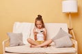 Unhappy sad little girl wearing casual clothing sitting on sofa at home against beige wall playing online video games on Royalty Free Stock Photo