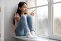 Unhappy school-aged korean girl sittng on windowsill at home Royalty Free Stock Photo