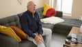 Unhappy senior man suffering knee pain, sitting alone on sofa indoor at home, sporting grey hair, glasses and worry expression Royalty Free Stock Photo