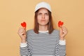 Unhappy serious woman wearing striped shirt and baseball cap standing isolated over beige background showing two little red hears