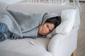 Unhappy sickly young woman sneezes covering face with hands lies on sofa wrapped in warm blanket