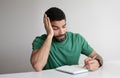 Unhappy tired pensive millennial arab man in green t-shirt make notes at notebook, planning startup