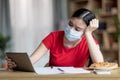 Unhappy tired pensive teenage asian girl in protective mask looking at tablet at table in room interior