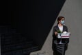 Unhappy unemployed woman in a protective mask is standing with a box of personal belongings against a gray wall