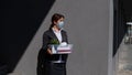 Unhappy unemployed woman in a protective mask is standing with a box of personal belongings against a gray wall
