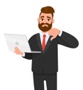 Unhappy young bearded business man holding/showing a latest new laptop and gesturing/making thumbs down sign with hand fingers.
