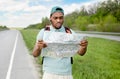 Unhappy young black man hitchhiking on road, looking at map, feeling lost, traveling alone by autostop