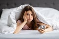 Unhappy young Caucasian woman lying in bed with alarm clock, not willing to get up in morning Royalty Free Stock Photo