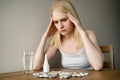 Unhappy young woman sitting at kitchen table among a lot of medicines