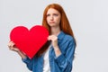 Unhapy brokenhearted cute redhead girl, sulking and looking up uneasy, distressed holding big red valentines day heart Royalty Free Stock Photo