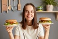 Unhealthy eating. Beautiful positive girl smiling at camera and posing with two tasty hamburgers, young pretty woman wearing in Royalty Free Stock Photo