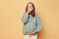 Unhealthy pregnant woman wearing knitted warm sweater standing isolated over beige background felling nausea covering mouth with Royalty Free Stock Photo