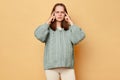 Unhealthy pregnant woman wearing warm knitted sweater touching painful temples posing over beige background suffering terrible