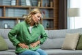 Unhealthy senior woman sitting on sofa at home. holding her stomach, grimacing in pain. She experiences severe abdominal