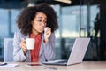 Unhealthy woman in formalware wiping face with napkin and holding white cup in office interior. Hispanic lady looking at