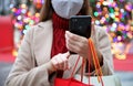 Unidentified woman with protective mask using mobile phone for shopping online and carrying bags on Christmas time. Focus on the