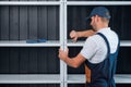 A uniformed worker is assembling a white metal shelving against a black clapboard wall