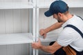 A uniformed worker staples white metal shelving to each other, against a white wall for storage in a warehouse or storeroom