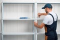 A uniformed worker staples white metal shelving to each other, against a white wall for storage in a warehouse or storeroom