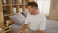 Unimpressed, the young hispanic man relaxes on his cozy bed, sitting comfortably with a serious face expression in his bedroom, Royalty Free Stock Photo