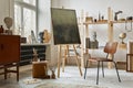 Unique artist workspace interior with stylish teak commode, wooden easel, bookcase, artworks, painting accessories, decoration.