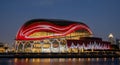 A unique modern building with an illuminated dome and a red and white pattern is seen at night by the waterfront with a city Royalty Free Stock Photo
