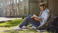 University student sitting under tree reading book with gripping plot, engrossed Royalty Free Stock Photo