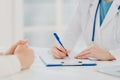 Unknown female doctor writes down prescription on papers in clipboard, consults patient about curing disease, pose at desktop