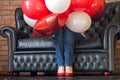 Unknown girl hidden in red and white helium balloons on sofa. Colorful balloons and women legs in red shoes. Birthday decoration.