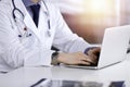 Unknown male doctor sitting and working with laptop in a darkened clinic, glare of light on the background, close-upof