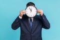 Unknown male person wearing official style suit hiding face behind wall clock display, wasting his
