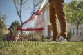 Unknown young man in casual clothes is using red garden rake on a lawn of his backyard. Useful tool of modern gardener