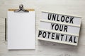 `Unlock your potential` on a lightbox, clipboard with blank sheet of paper on a white wooden background, top view. Flat lay, Royalty Free Stock Photo