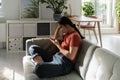 Unmotivated confused Asian woman sits with phone on sofa in apartment not willing to start working