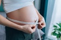 unrecognisable pregnant woman measuring her growing abdomen size with measuring tape at home.