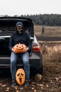 An unrecognizable adult man wearing a skull mask sits in the trunk of a car with carved pumpkins for Halloween.