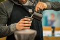 Unrecognizable Barista wearing black apron making cappuccino, pouring milk in paper cup in coffee bar