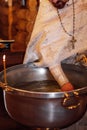 Unrecognizable bearded priest in silver cassock stand at metal font, consecrating water for ceremony of infant baptism.