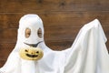 Unrecognizable boy in white costume of halloween smiling ghost holds little decorated pumpkin, on wooden background ready for cele