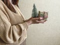 An unrecognizable brunette girl in a beige sweater holds a Christmas gift in eco packaging and a green Christmas tree in her hands Royalty Free Stock Photo
