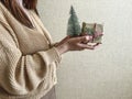 An unrecognizable brunette girl in a beige sweater holds a Christmas gift in eco packaging and a green Christmas tree in her hands Royalty Free Stock Photo