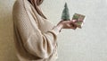 An unrecognizable brunette girl in a beige sweater holds a Christmas gift in eco packaging and a green Christmas tree in her hands Royalty Free Stock Photo