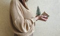 An unrecognizable brunette girl in a beige sweater holds a Christmas gift in eco packaging and a green Christmas tree in her hands Royalty Free Stock Photo