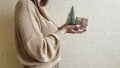 An unrecognizable brunette girl in a beige sweater holds a Christmas gift in eco packaging and a green Christmas tree in her hands Royalty Free Stock Photo